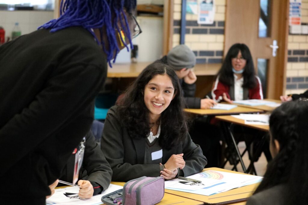 A woman talking to a female pupil sitting at a desk in a classroom